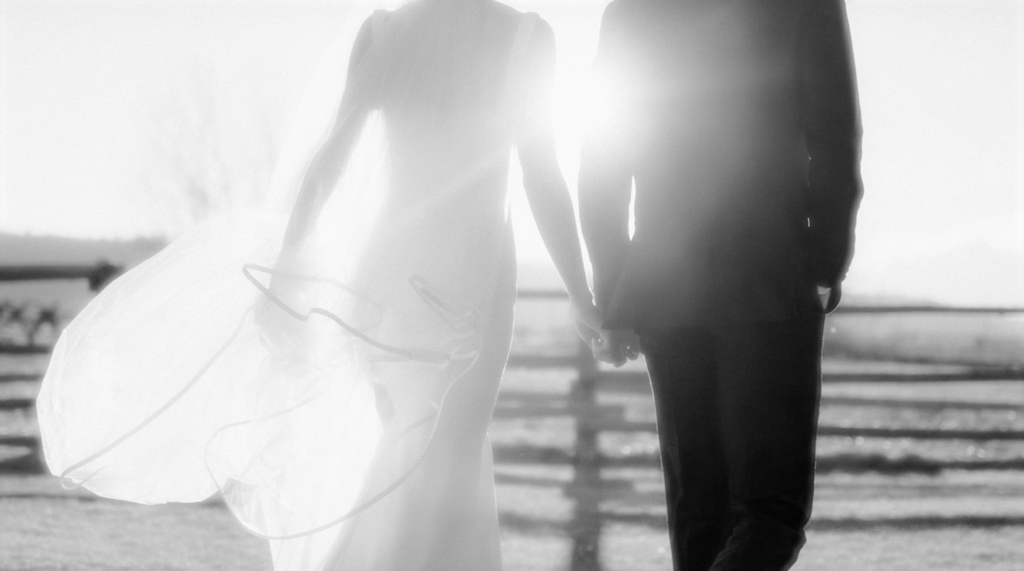 Bride and groom hold hands walking along a wooden fence.